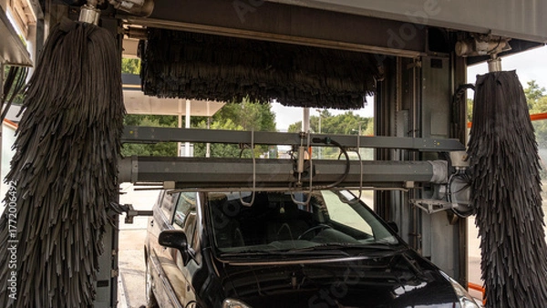 Fototapeta Front view of a dark car inside an automatic car wash. The large side brushes and the upper brush of the machine are visible, ready to clean the vehicle.