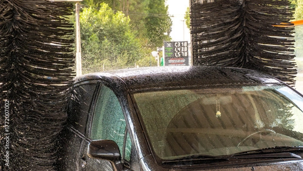Obraz Front view of a dark car inside an automatic car wash. The large side brushes and the upper brush of the machine are visible, ready to clean the vehicle.