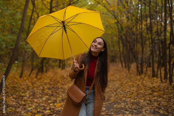 Fototapeta Happy young woman with long hair holding yellow umbrella, walking in autumn park surrounded by golden trees and fallen leaves, smiling and looking up at the sky.