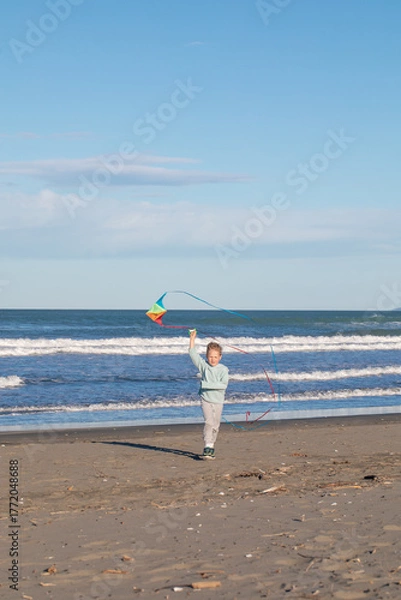 Fototapeta Joyful young boy running along the beach and flying a colorful kite. Fun outdoor activity for kids