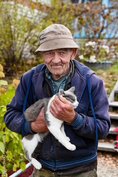 Fototapeta Close up portrait of elderly Russian man in autumn garden holding his gray and white cat with affection, peaceful rural lifestyle in Russia.