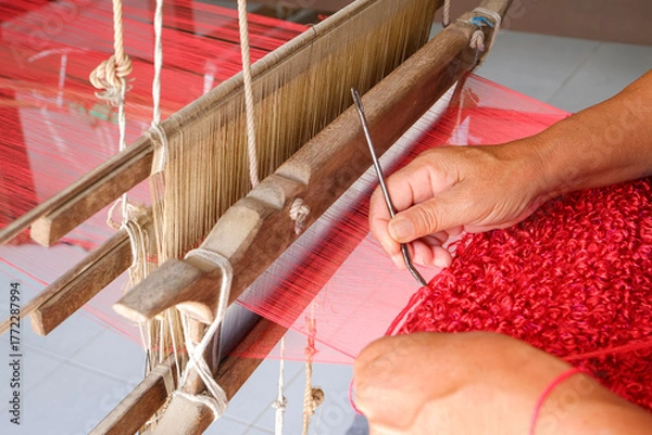 Fototapeta Close up of hands weaving red silk fabric texture pattern with needle on a traditional wooden loom, showing local craftsmanship, cultural heritage, and handmade textile art in Thailand.