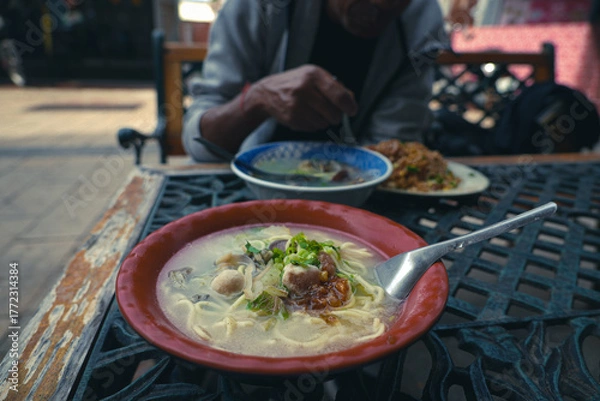 Fototapeta Close-up of a seafood soup noodles bowl on the table with a cropped image of a man eating fried rice in the blur background. 