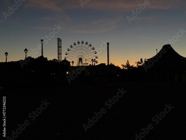 Fototapeta Evening view of amusement park at sunset with ferris wheel silhouette.