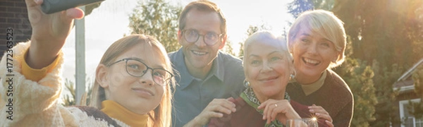 Fototapeta Header of teenage Caucasian girl taking selfie with middle aged Caucasian man, senior Caucasian woman, middle aged Caucasian woman smiling outdoors, all looking at smartphone camera
