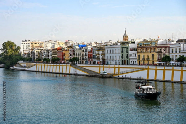 Obraz Colourful riverside buildings of the Triana district reflected on the Guadalquivir River in Seville, with a boat gliding calmly through the water