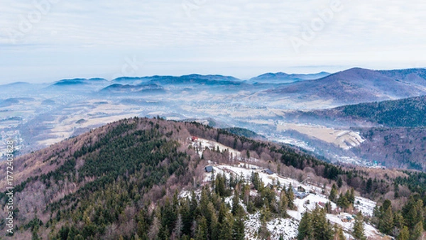 Obraz A tranquil winter panorama of Beskid Mały in southern Poland, where snowy meadows meet evergreen forests and rolling mountain hills under a serene, cloud-streaked sky.