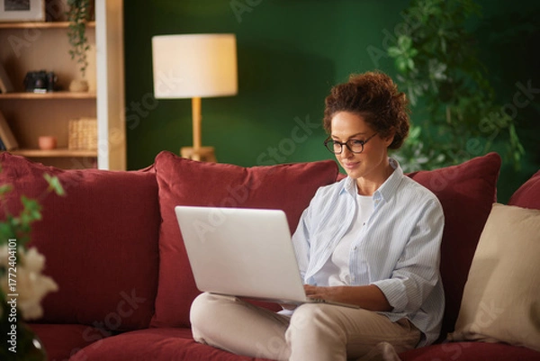 Fototapeta A woman sits on a red couch in a bright living room, focused on her laptop. She wears glasses and a casual shirt, creating a relaxed atmosphere in the afternoon light.