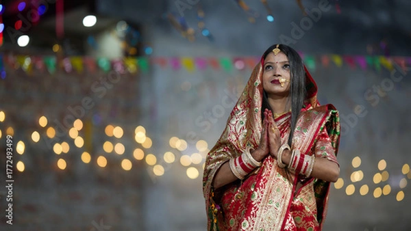 Fototapeta Newly married Indian woman in red saree doing namaskar with diya lights and bokeh festival background, symbolizing devotion and grace.