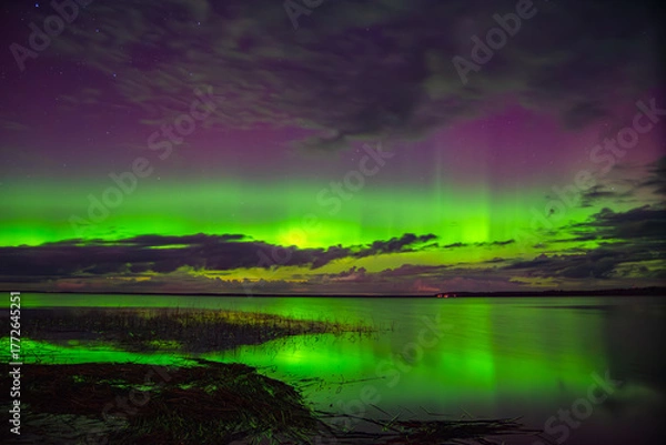 Obraz Vivid northern lights glowing green and purple over a calm lake with reflections and scattered clouds under a starry night sky.
