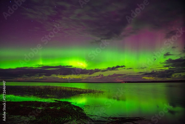 Obraz Vivid northern lights glowing green and purple over a calm lake with reflections and scattered clouds under a starry night sky.