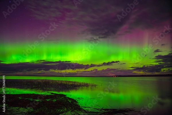 Obraz Vivid northern lights glowing green and purple over a calm lake with reflections and scattered clouds under a starry night sky.