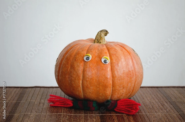 Fototapeta Yellow ripe pumpkin with perfect shape, with a scarf on Halloween and eyes, on a wooden table on a white background.