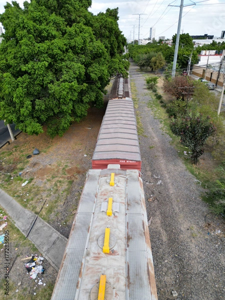 Fototapeta Vertical aerial drone shot showcasing a long freight train on railway tracks in Guadalajara, Mexico. Lush green trees line the urban route, with a glimpse of city infrastructure and a parallel road