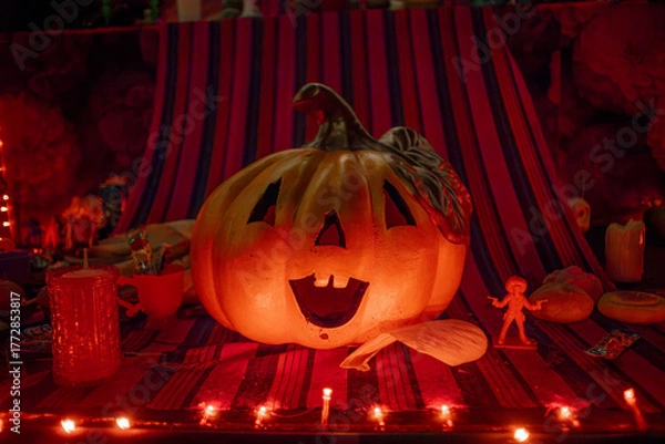 Fototapeta Illuminated carved pumpkin Jack-o'-lantern on a festive Day of the Dead altar or display, with atmospheric red lights.