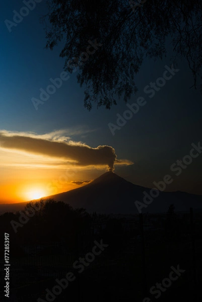 Fototapeta Mystical silhouette of Popocatépetl volcano erupting at sunrise. Fumarole over the peak, framed by tree branches
