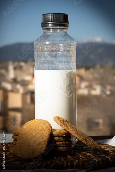 Obraz Botella de leche y galletas integrales sobre una mesa al aire libre con un fondo de paisaje mediterráneo y cielo azul. 