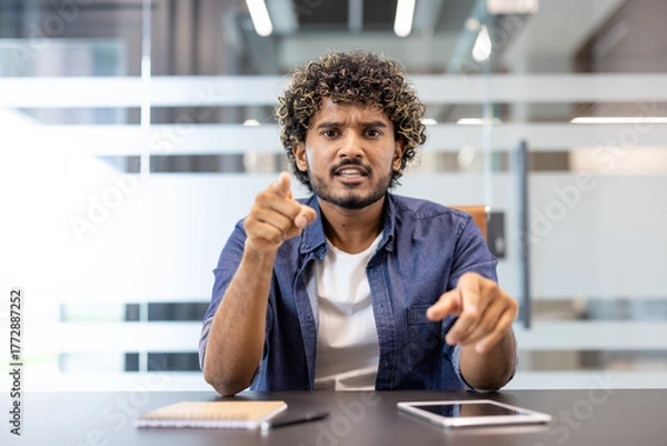 Fototapeta Portrait of a young angry Indian man sitting in the office facing the camera and talking emotionally