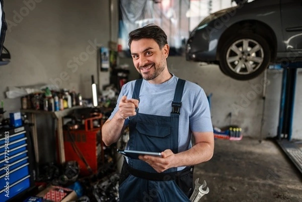 Fototapeta Confident mechanic pointing at camera and holding digital tablet in auto repair shop