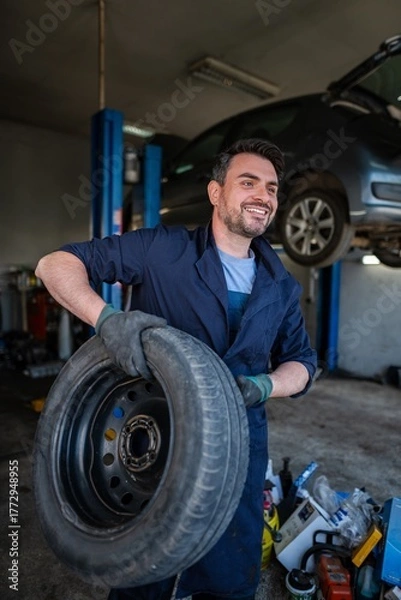 Fototapeta Mechanic holding a tire and smiling in a car repair shop