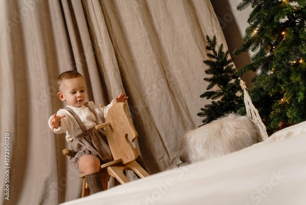 Obraz Little boy in casual overalls and socks rides a rocking horse near a Christmas tree