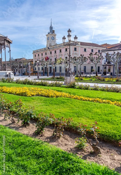 Obraz The pink-hued, clock-towered Alcalá City Hall provides a backdrop to the Plaza de Cervantes's formal gardens, featuring rows of yellow flowers, green grass, and pollarded plane trees