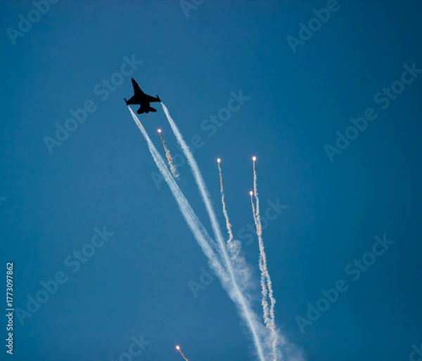 Fototapeta A fighter plane is seen throwing heat flares (flare) in the blue sky background. Bright light traces and smoke clouds reveal the dynamism and visual appeal of the maneuver.