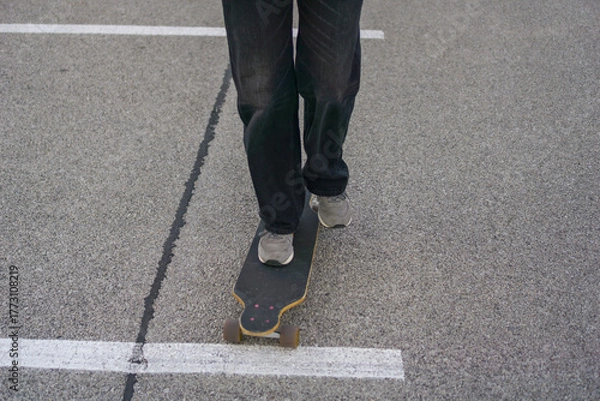 Fototapeta low angle close up of a persons feet in gray sneakers standing on a dark longboard on asphalt, white parking space line running under the board, concept of action sports