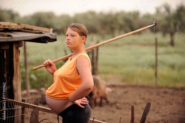 Fototapeta A pregnant girl in an orange blouse arranges order on the plot of land on the estate with a pig paddock on a hot summer day