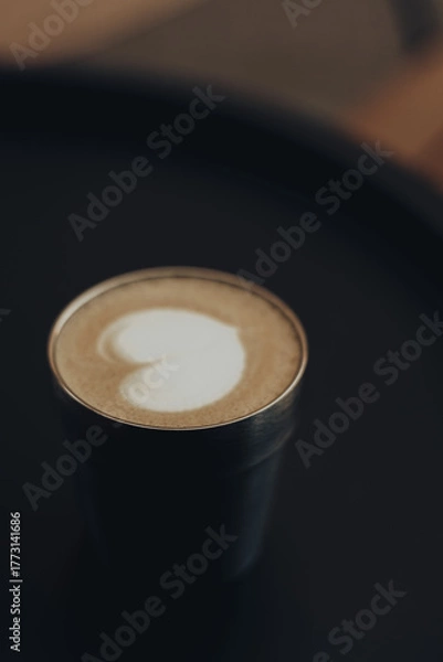 Fototapeta Minimalist coffee aesthetic with creamy latte art in a stylish metal cup on a dark background. Warm tones, soft focus and elegant composition create a cozy, modern cafe vibe. Coffee shop. Hot drink.
