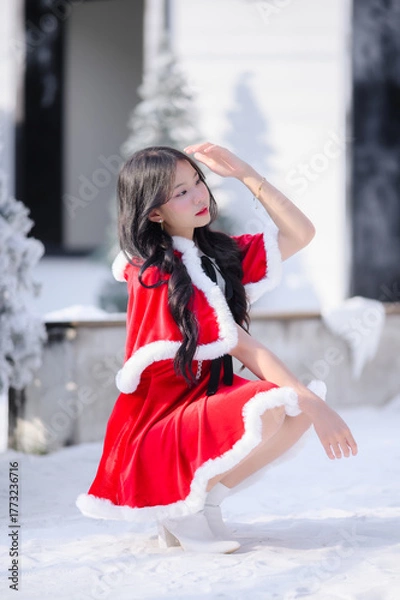 Obraz young asian woman wearing a red santa dress with white fur trim smiling and posing outdoors in a snowy Christmas scene, surrounded by frosted pine trees and bright winter sunlight