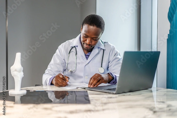 Obraz Young doctor man working at office workplace, reading medical record, paper document at laptop, reviewing patient history, sitting at table with laptop computer, working in clinic office