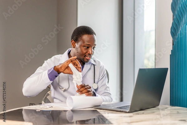 Obraz Male doctor is explaining something using an anatomical model bone while having online consultation with a patient, talking scoliosis, herniated discs, osteoporosis, diagnosing bone density issues