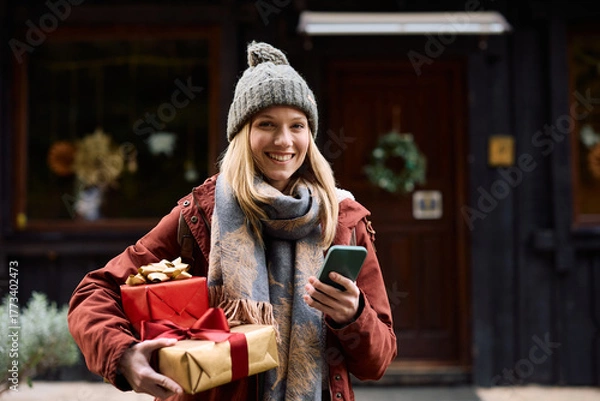 Fototapeta Happy woman using cell phone while holding Christmas presents on front of her house.