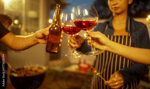 Obraz An Asian woman and friends toast with wine during a cozy backyard BBQ. Grilling skewers over charcoal under string lights, they celebrate friendship, joy,  togetherness in a festive outdoor evening