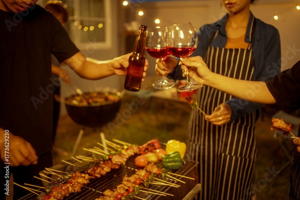Fototapeta An Asian woman and friends toast with wine during a cozy backyard BBQ. Grilling skewers over charcoal under string lights, they celebrate friendship, joy,  togetherness in a festive outdoor evening
