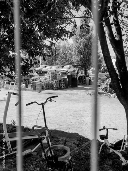 Obraz A black and white view through wire fencing, showing a cluttered landfill site with stacked pallets, old equipment, bicycles, and cars mounted on a metal structure for attention