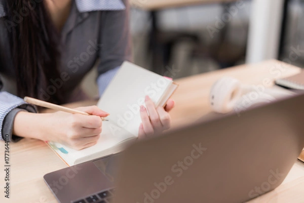 Fototapeta Woman's hands writing notes in an open notebook with a pencil, focusing on tasks while using a laptop and headphones on a desk, representing online learning and remote work concepts