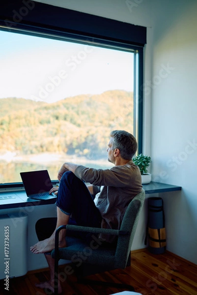 Fototapeta Man sitting barefoot at a desk, looking out a large window at a scenic landscape, a laptop open next to him