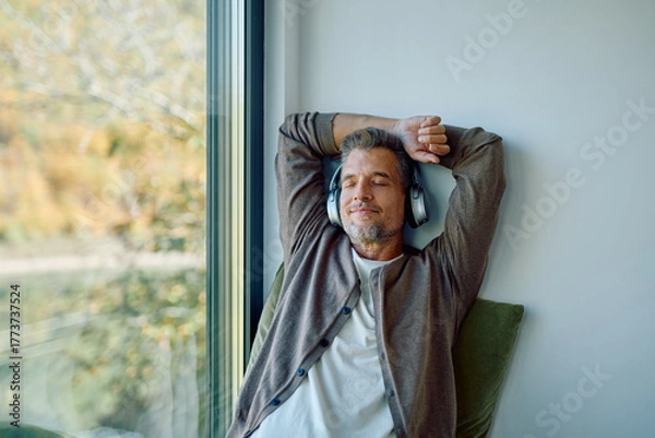 Fototapeta Happy man wearing headphones, eyes closed, enjoying calm music and relaxation by the window in his cozy home
