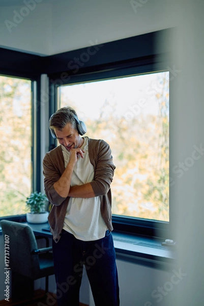Fototapeta Man with headphones listening to music, having a relaxed moment near a modern window, showing happiness and contemplation
