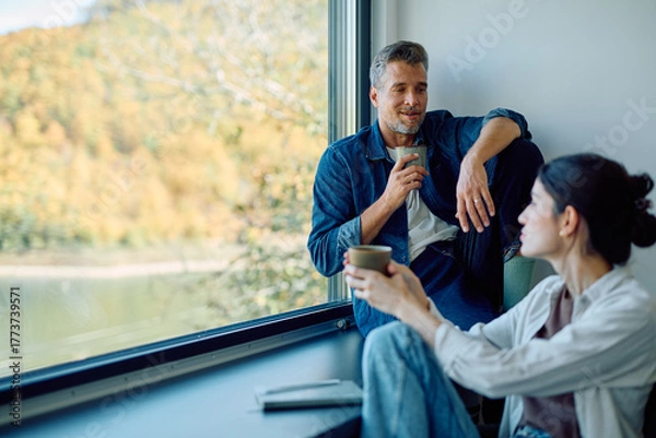 Fototapeta Happy adult couple enjoying coffee and conversation, sitting by a large window with autumn nature views