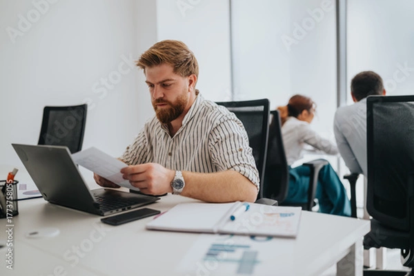 Obraz A focused office worker sits at a clean desk, examining papers beside a laptop. The scene conveys professionalism, productivity, and a modern, collaborative workspace.