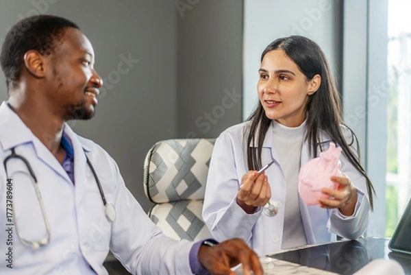 Fototapeta Two cardiologists engage in a detailed discussion on heart disease treatment, using an anatomical model to illustrate their points while consulting with a medical intern in a clinic, share knowledge