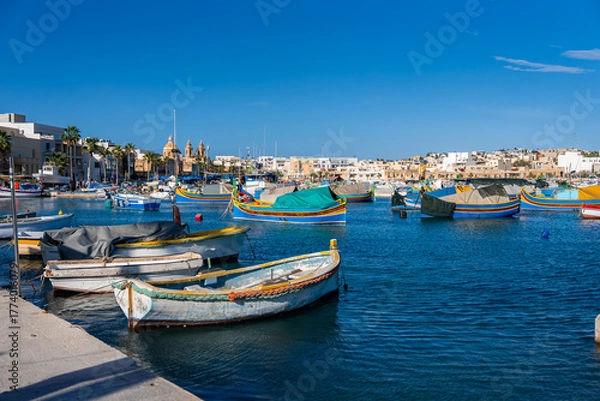 Fototapeta Brightly painted luzzu boats bob in Marsaxlokk, Malta, as reflections dance on deep blue water. The church dome and twin bell facade rise beyond limestone quays on a clear day.