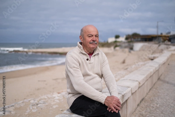 Fototapeta Elderly man with a mustache sitting on a seawall by the ocean 
