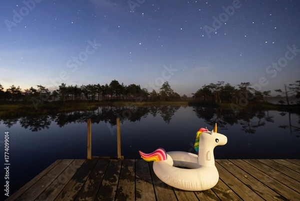 Fototapeta Inflatable unicorn float on a wooden dock under a clear, starry night sky reflected in a calm dark lake.