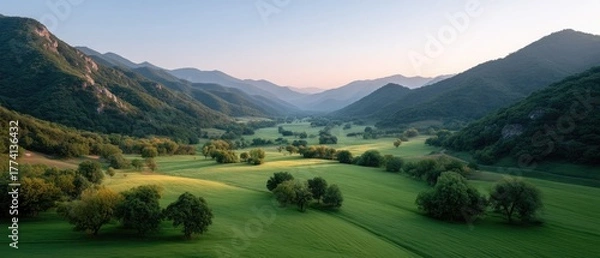 Obraz Lush Green Valley Illuminated by Golden Sunlight During a Serene Sunrise Over Rolling Hills and Distant Mountains