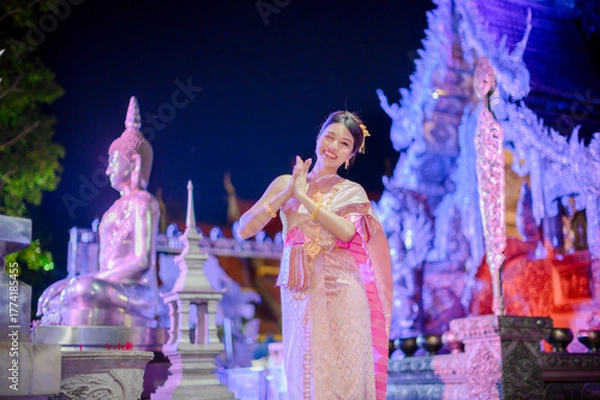 Obraz young woman wearing a traditional thai dress smiling and greeting with wai gesture in front of wat sri suphan temple during yi peng and loy krathong festival night