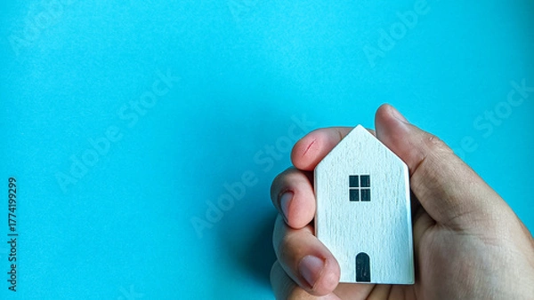 Obraz Minimalist concept image showing a hand gently holding a small wooden house on a blue background, symbolizing home ownership, safety, real estate, family care, and housing security.
